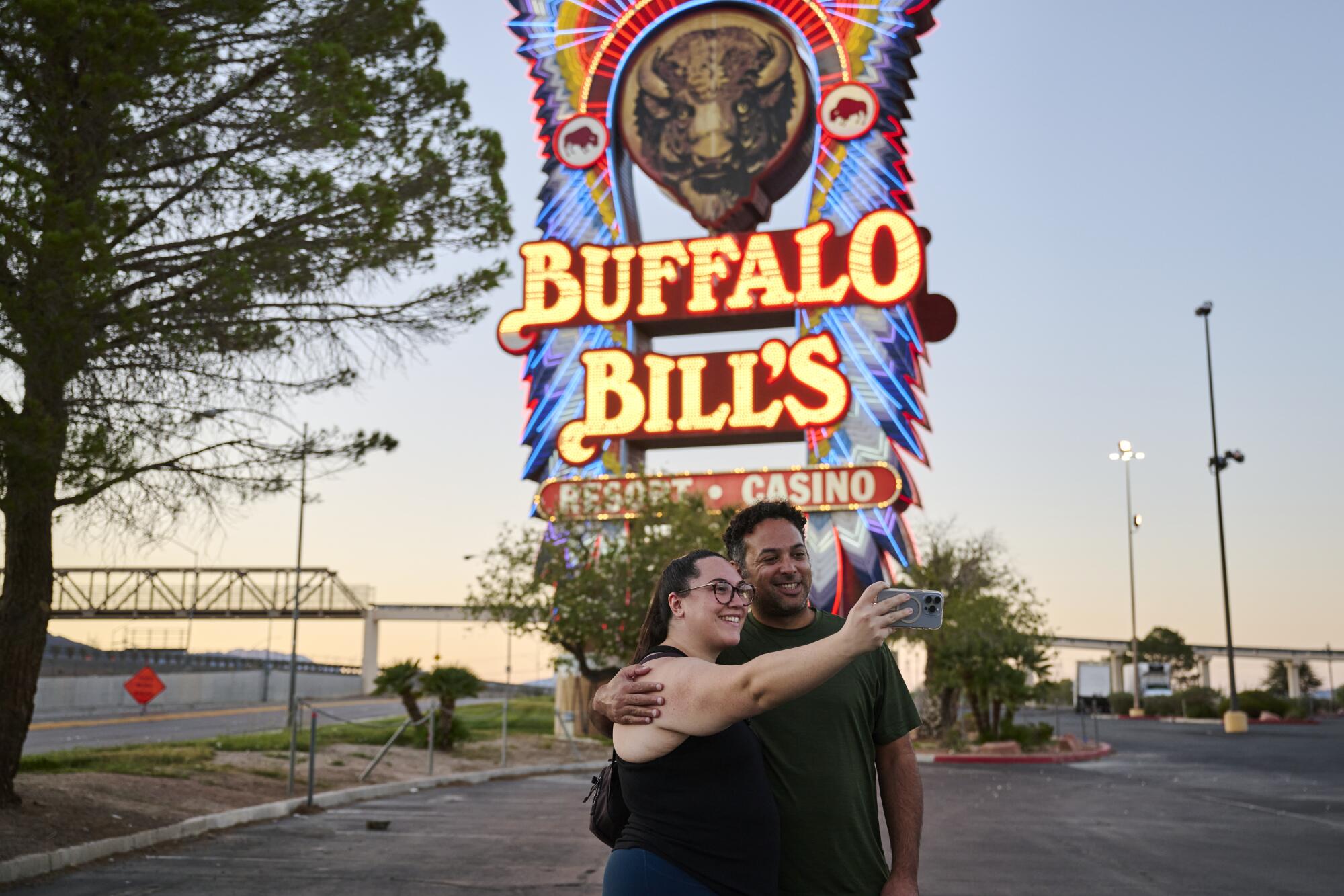 A man and woman pose for a photo in an empty parking lot in front of a neon sign at sunset.