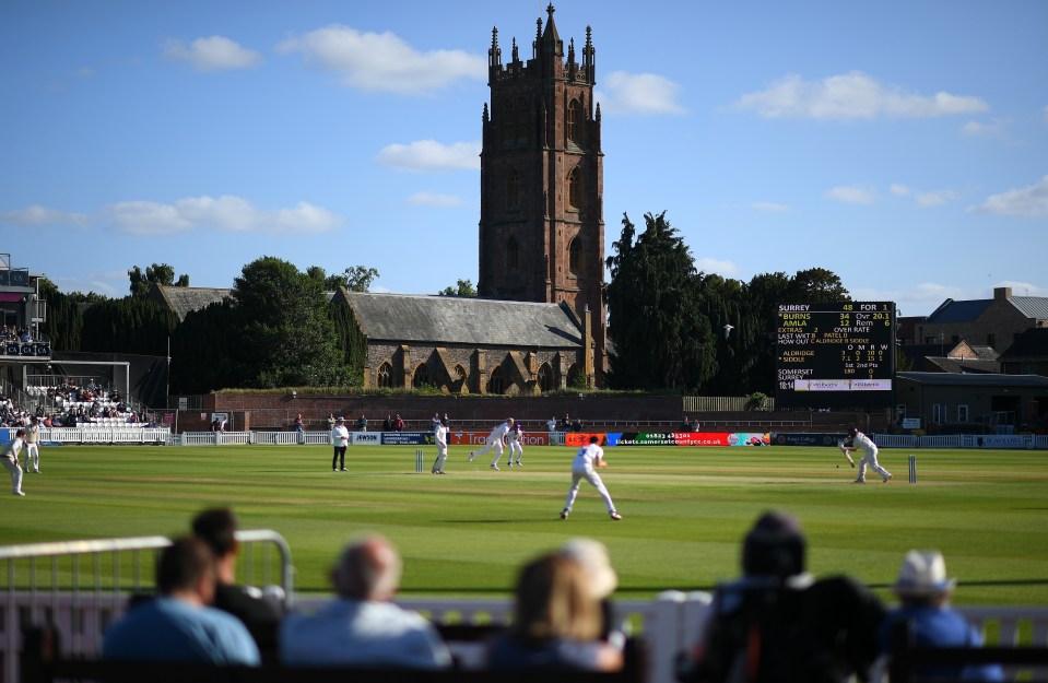 General view of a cricket match between Somerset and Surrey, with a large scoreboard and a historic church tower in the background.
