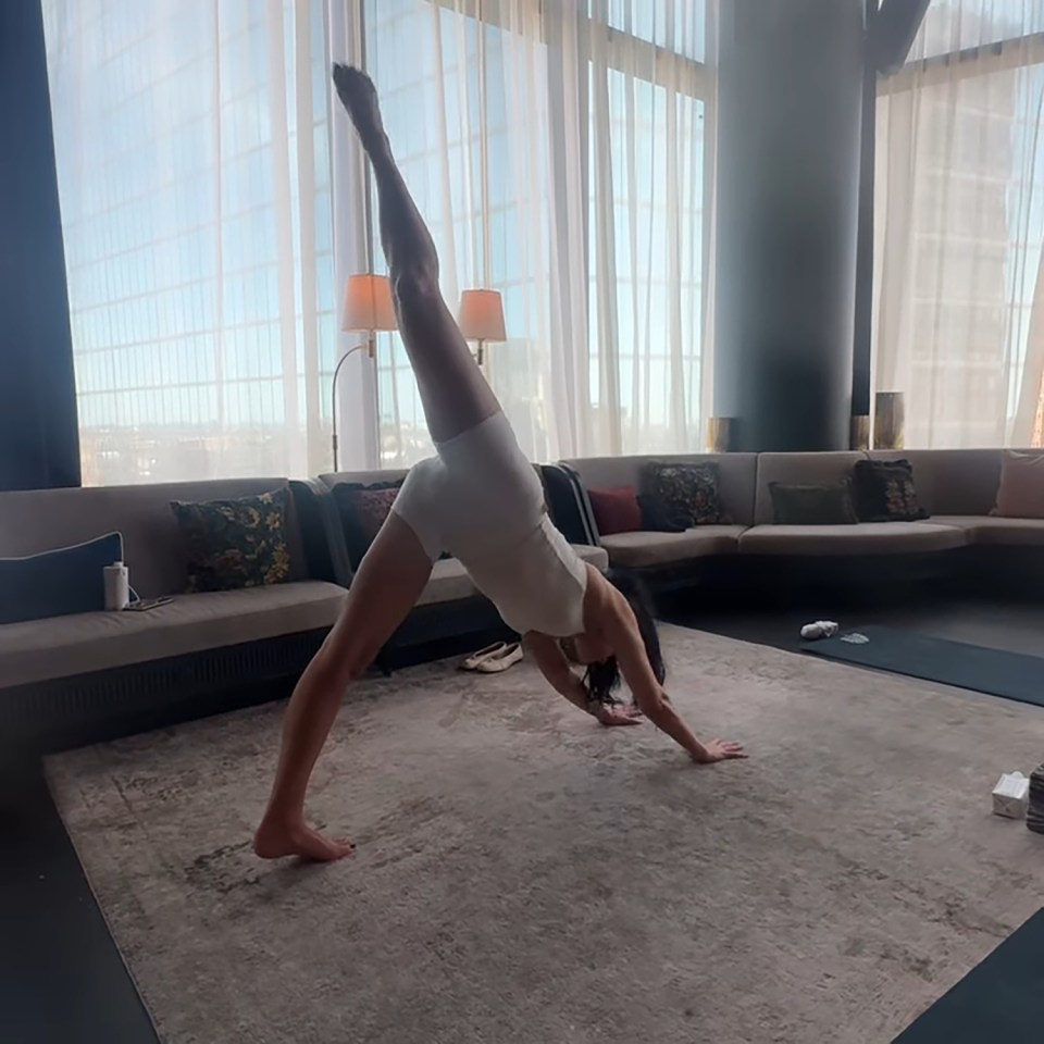 Dua Lipa in a white leotard performing a yoga pose, lifting one leg straight into the air in a downward dog variation.