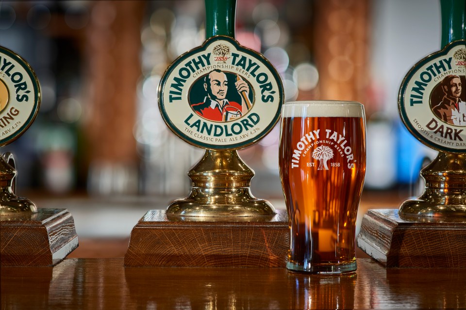 A glass of Timothy Taylor's beer next to a beer tap on a wooden bar.
