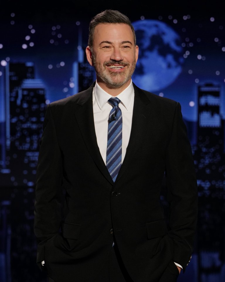 Jimmy Kimmel smiling, wearing a suit and striped tie, in front of a backdrop of a city skyline at night with a full moon.