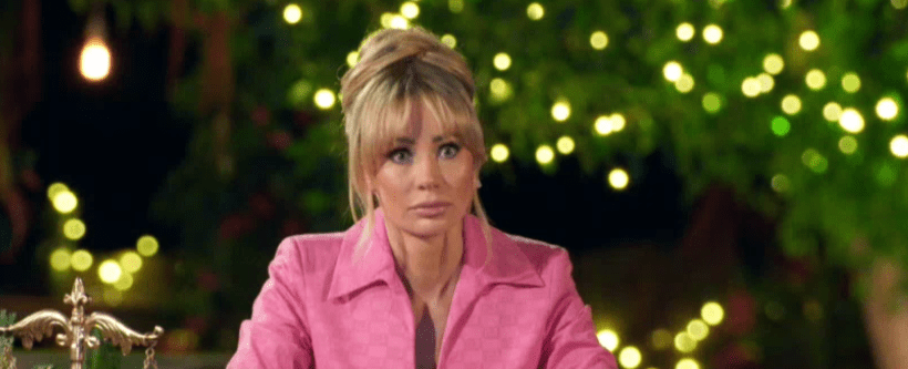 A woman with blonde hair, wearing a pink jacket, looking surprised against a blurred background of green foliage and fairy lights.