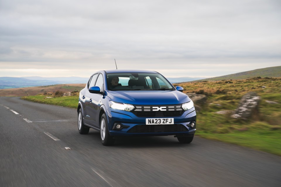 A blue Dacia Sandero hatchback driving on a rural road with hills in the background.