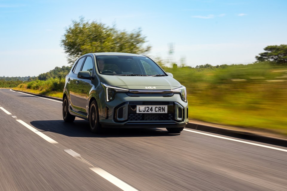 A green Kia car driving down a road with blurred green fields and trees in the background.