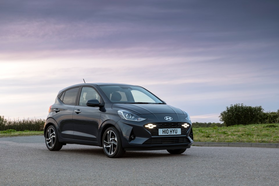 Dark gray Hyundai i10 parked on an asphalt road with a grassy area and bushes to the right and a pale sky with hints of pink and purple.