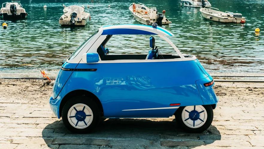 Blue and white Microlino Spiaggina EV next to a body of water with boats.