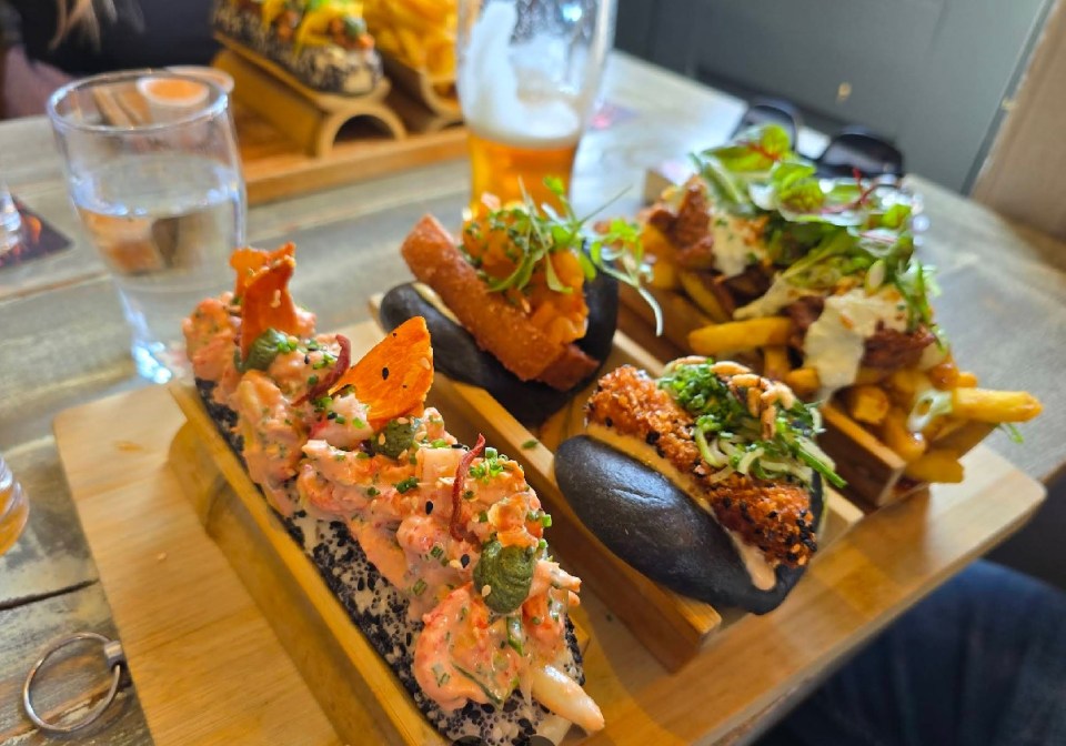 Variety of pub dishes on a wooden serving board, including a lobster roll, fried seafood buns, and loaded fries.