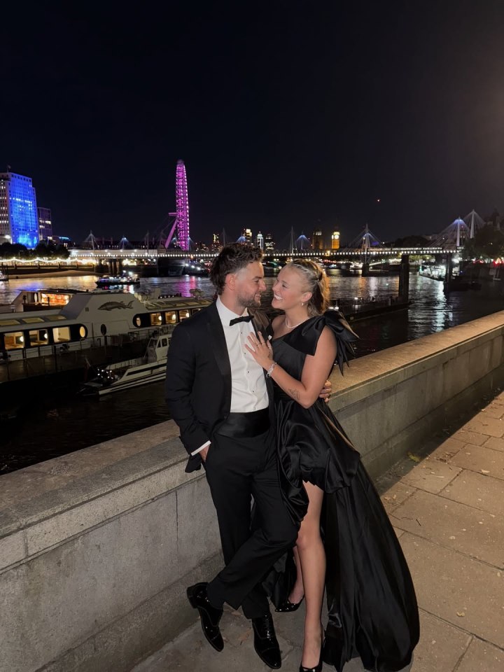 Jojo Siwa and Chris Hughes embracing by a river at night with city lights in the background.