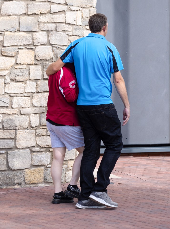 A person in a blue shirt with their arm around a person in a red jacket, walking away from a stone wall.