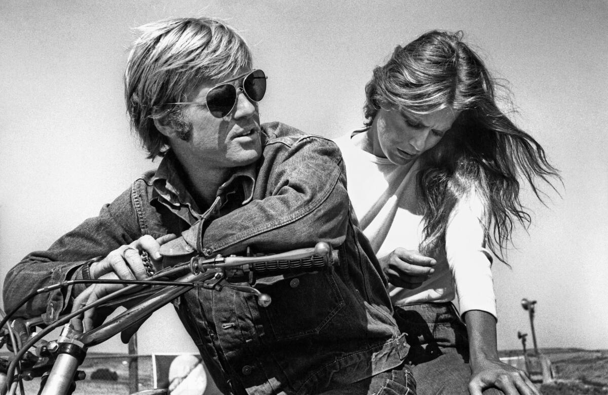 A man in shades sits on a motorcycle with a woman riding behind him.