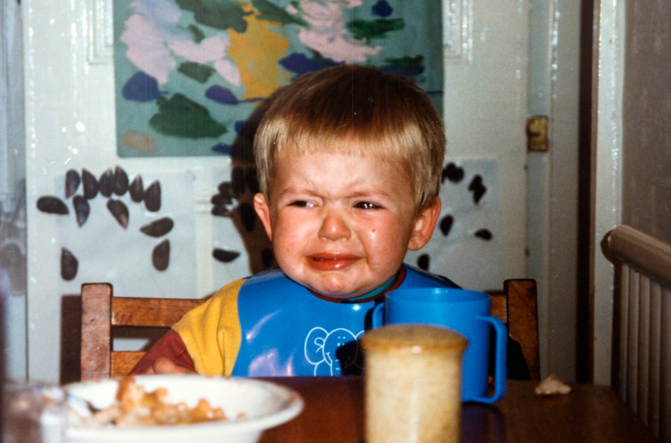 Andrew Gosden as a 2-year-old in a blue bib and yellow shirt, sitting at a table with a white bowl, crying.