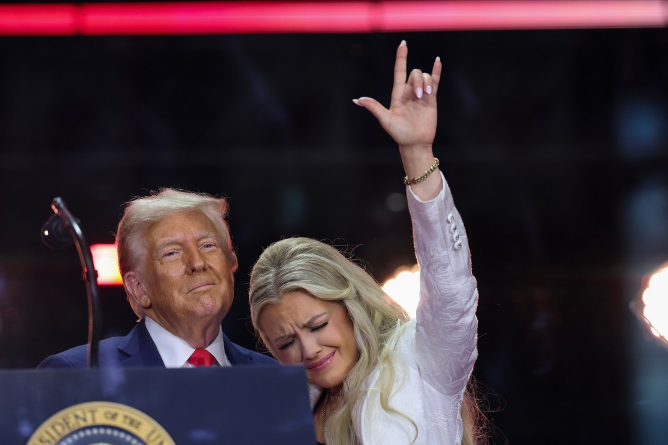 Erika Kirk crying onstage with her arm raised in a "rock on" gesture, with former President Donald Trump in the background at a memorial service.