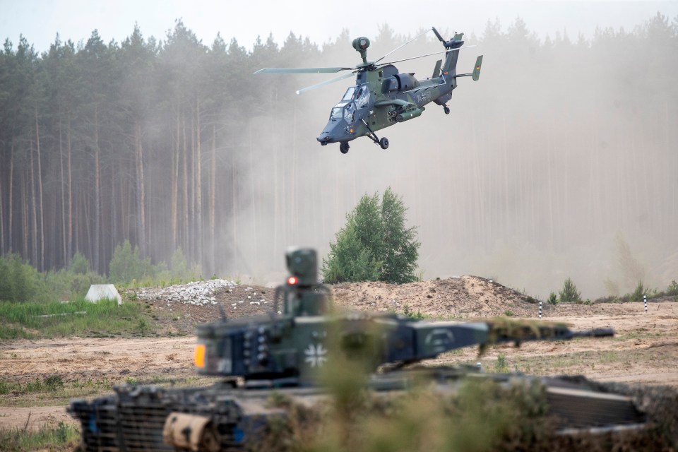 A German Army Eurocopter Tiger helicopter flying over a military training range in Lithuania, with a tank in the foreground.