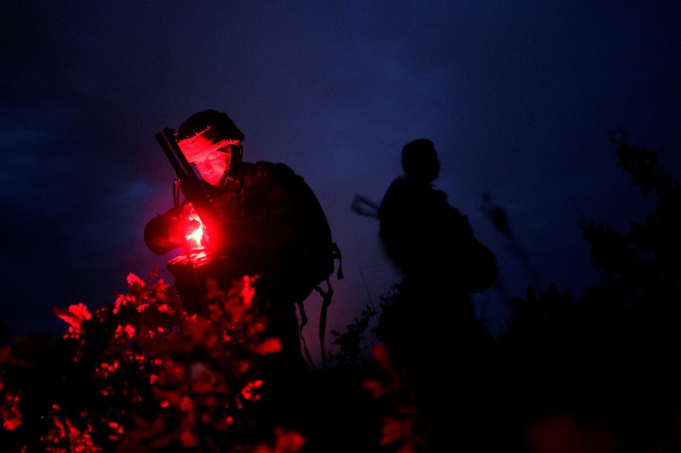 A Ukrainian soldier with a red light illuminating his face and rifle, silhouetted against a dark blue sky.