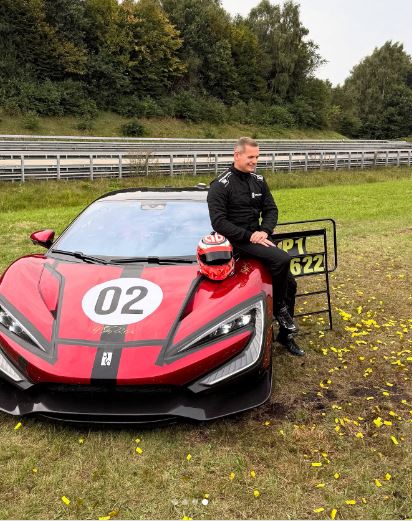 A man in a racing suit sitting on the red Yangwang U9 Xtreme car with a racing helmet on the hood.