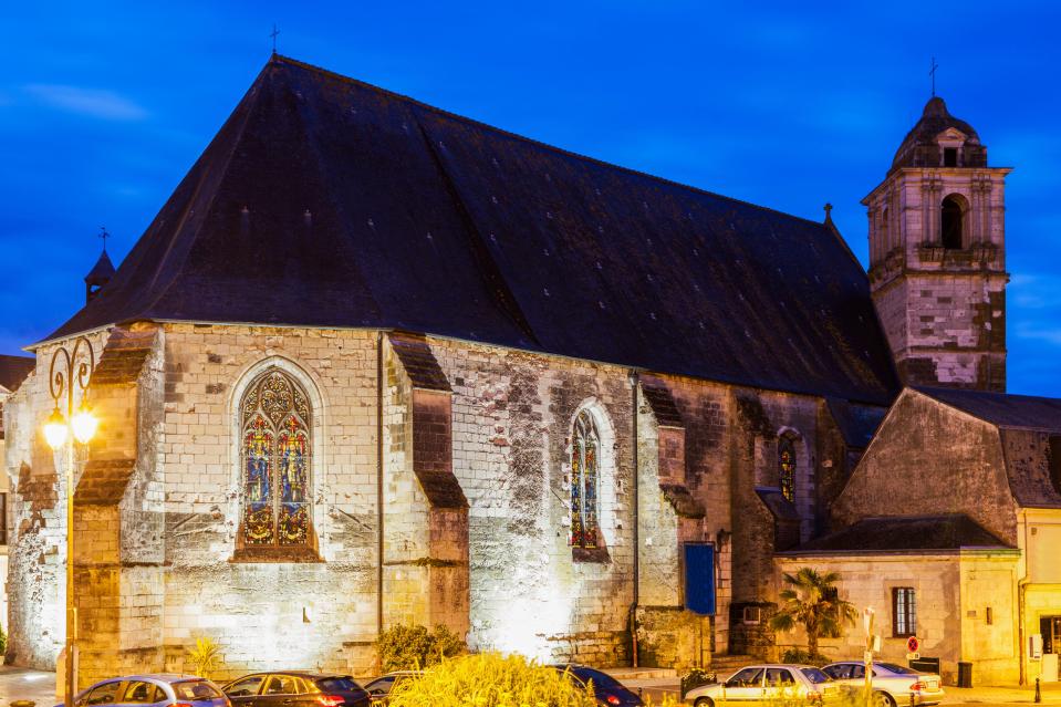St. Florentin Church in Amboise, France, at dusk, with its large, dark roof and illuminated stone walls featuring stained glass windows, and a tall bell tower rising in the background.