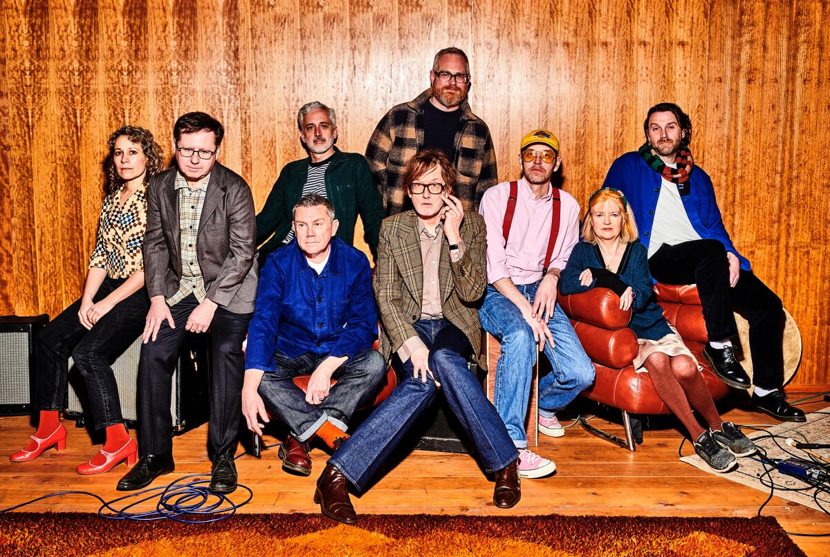 Several members of Pulp surround lead singer Jarvis Cocker, who wears a plaid blazer and blue jeans.