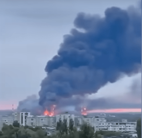 Large plume of dark smoke rising above a city with fires visible below.