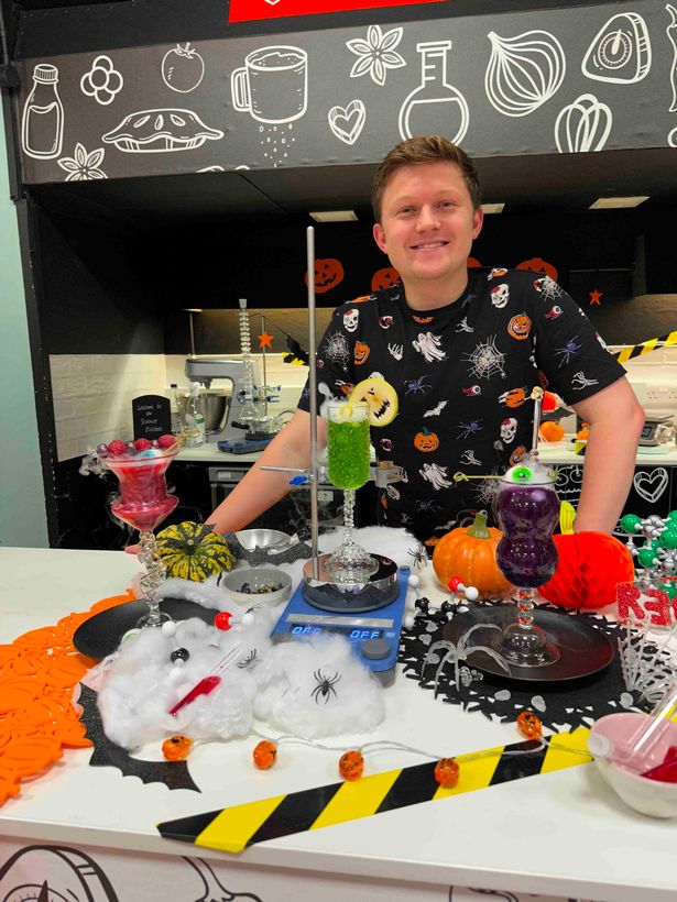 Josh Smalley, in a black top with Halloween symbols on, stood behind a desk, covered in decorations, in a science lab.