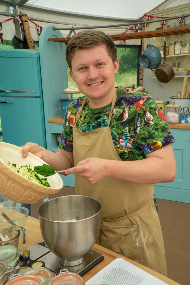Josh Smalley, in a green top and beige apron, baking in the Great British Bake Off tent.