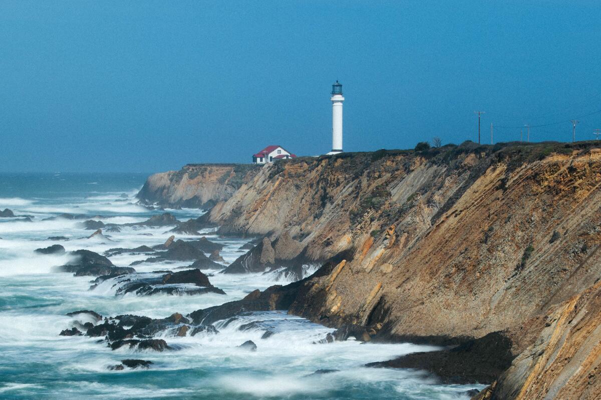 A white lighthouse tower is visible in the background; foreground is the rocky California coastline and blue crashing waves.