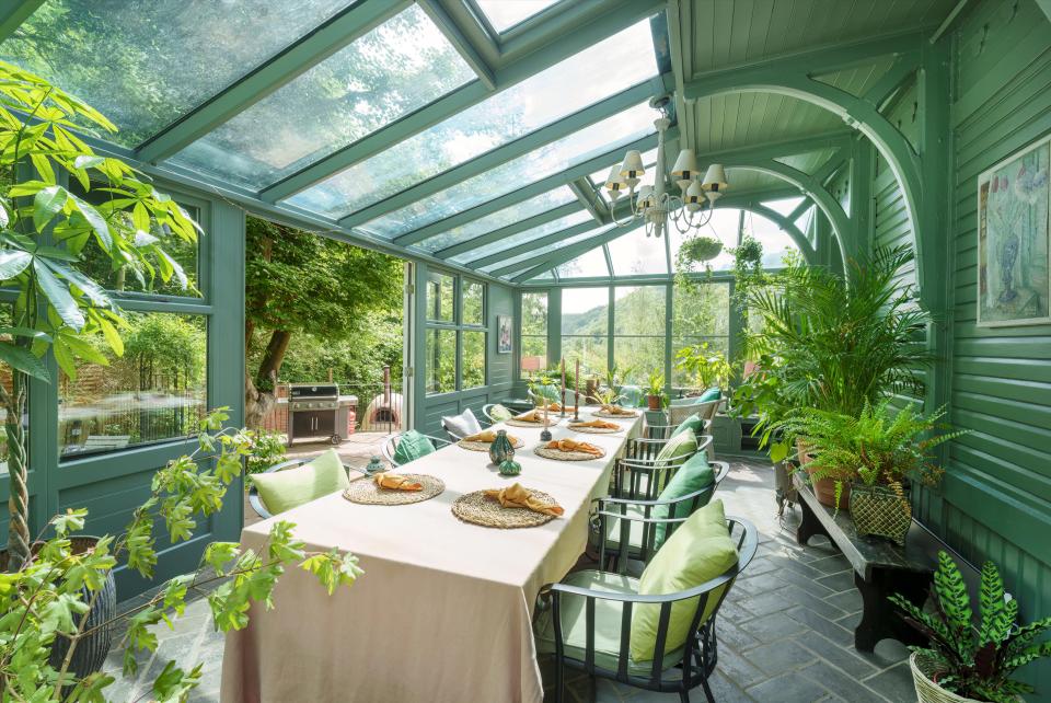 A dining room with green walls and glass ceilings, a long table set for dining, and many potted plants.