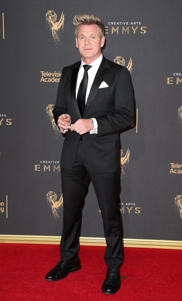 Gordon Ramsay in a black suit on a red carpet with Emmy logos in the background.