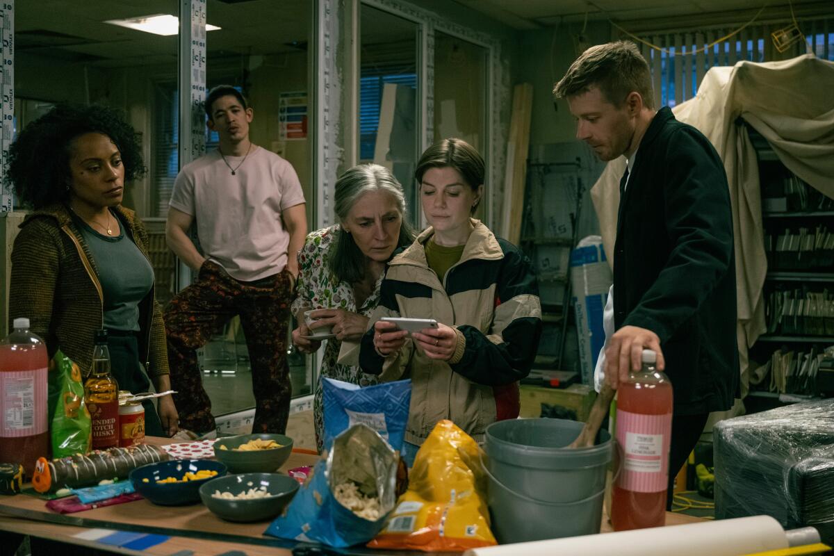 A group of men and women are gathered around a table and looking at a cell phone.