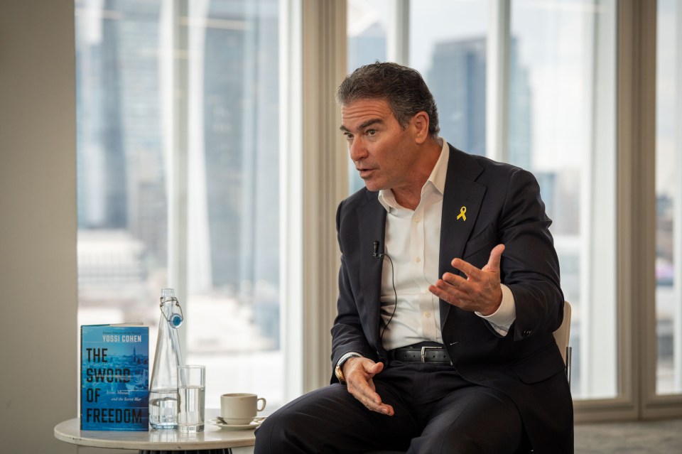 Yossi Cohen, former head of Mossad, in a suit with a yellow ribbon pin, speaking with a copy of "The Sword of Freedom" book next to him.