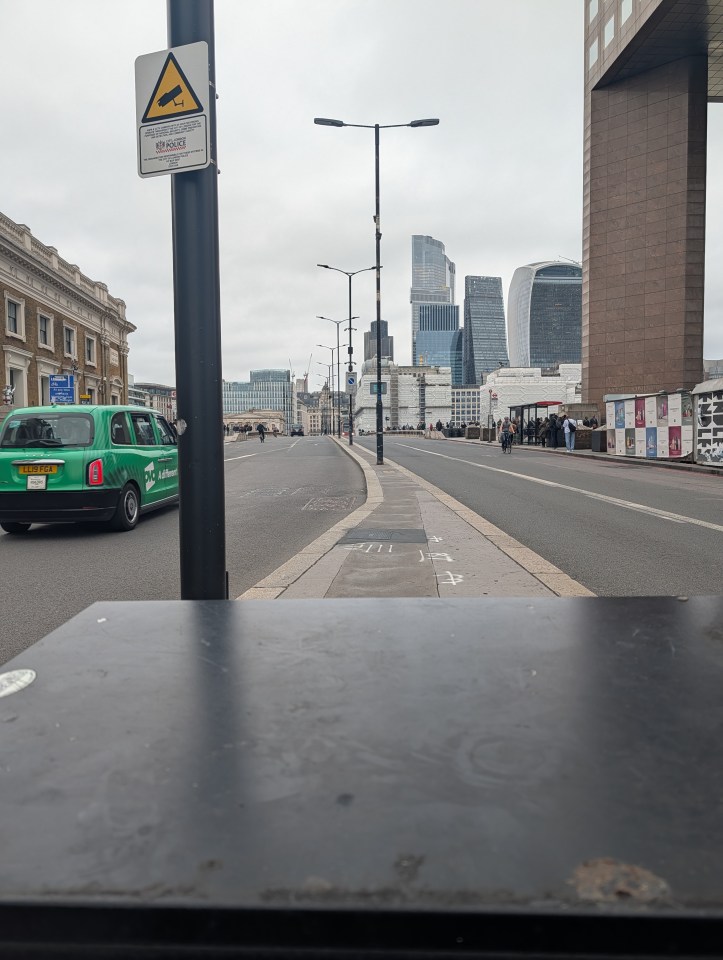 A street scene with a green taxi on the left, a pedestrian and bike lane on the right, and modern skyscrapers in the background under a cloudy sky.