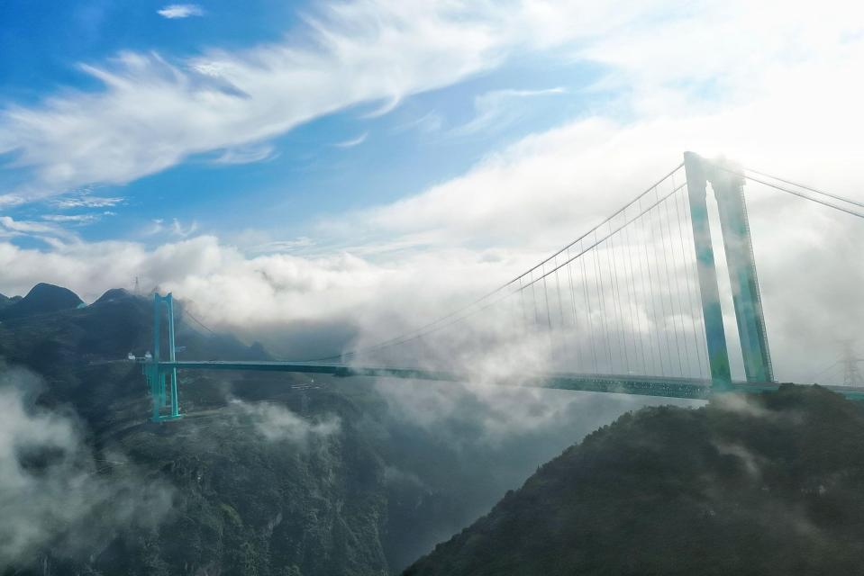 Aerial photo of the Huajiang Grand Canyon Bridge in southwest China's Guizhou Province, with clouds obscuring parts of the bridge and mountainous terrain.
