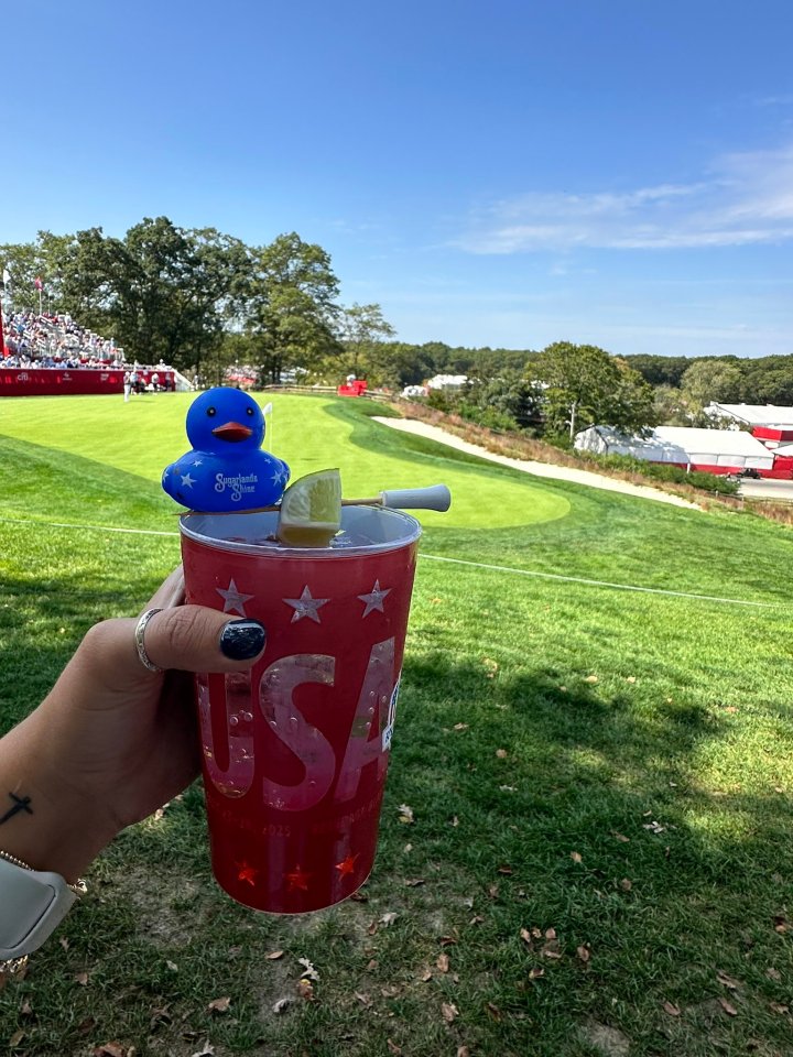 A red cup with "USA" printed on it and a blue rubber ducky in it is held up to a golf course, where spectators are in the background.