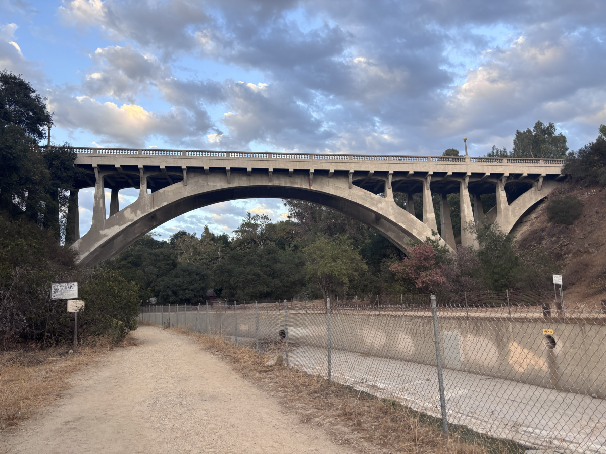 An open-spandrel reinforced concrete arch bridge above a dirt path and concrete river channel.