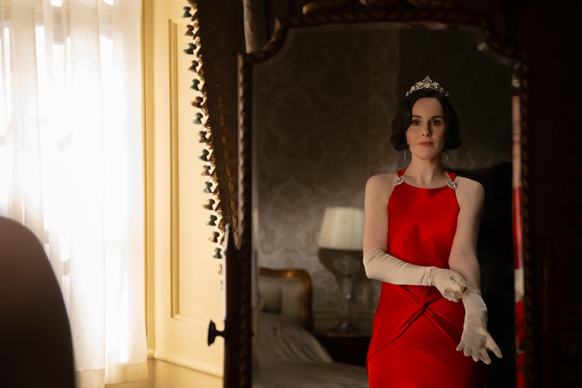 A woman in a red dress and a tiara preps in front of a mirror.