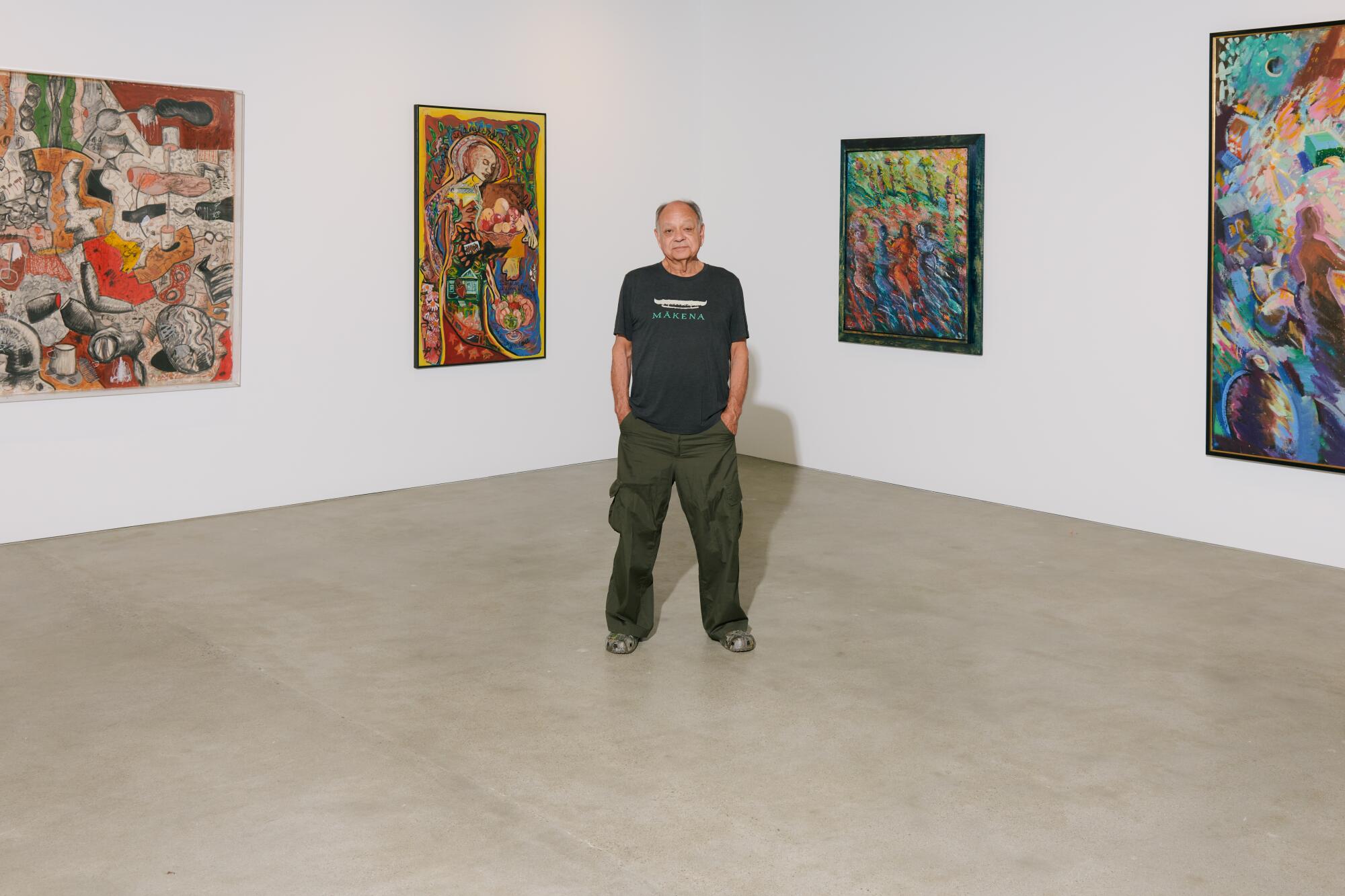 Cheech Marin, wearing a black t-shirt and cargo pants, stands between paintings in a gallery