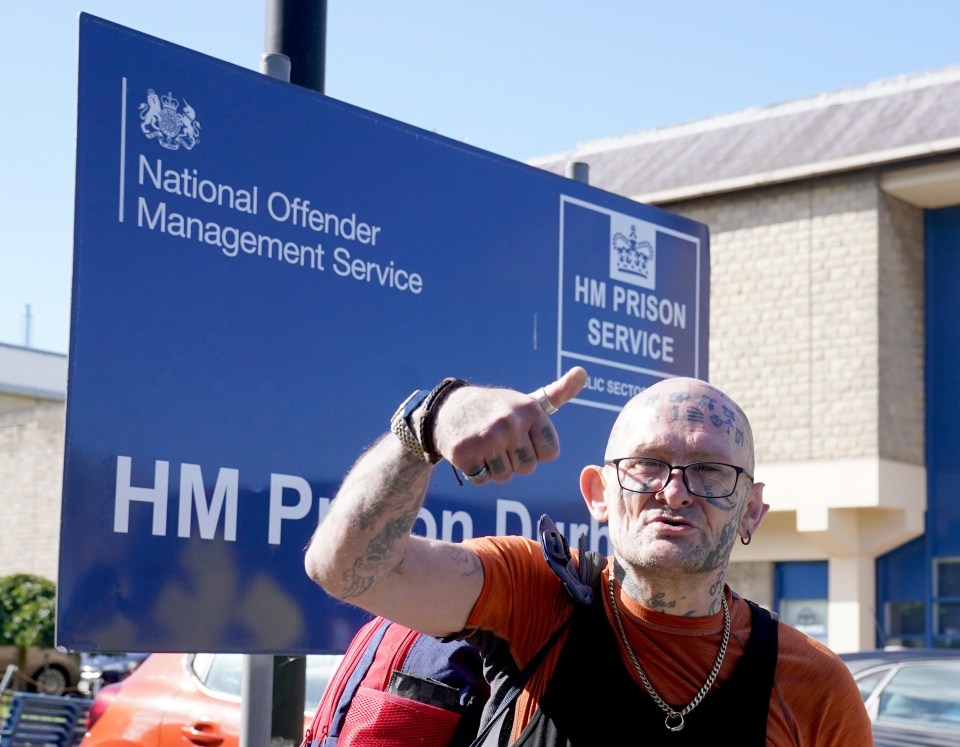 Jason Hoganson with multiple facial tattoos, wearing glasses, giving a thumbs-up sign in front of a blue sign for HM Prison Durham.