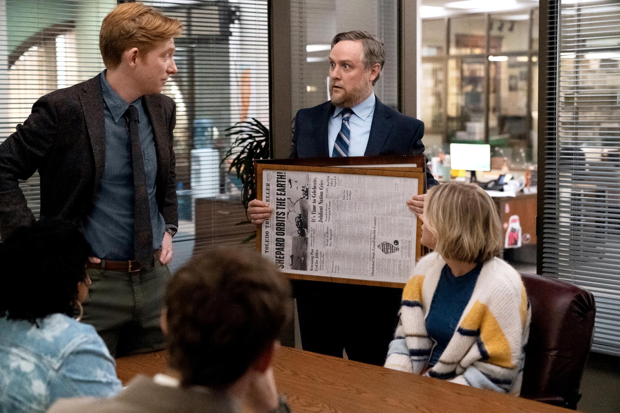 A man in a suit holds a framed newspaper in front of colleagues