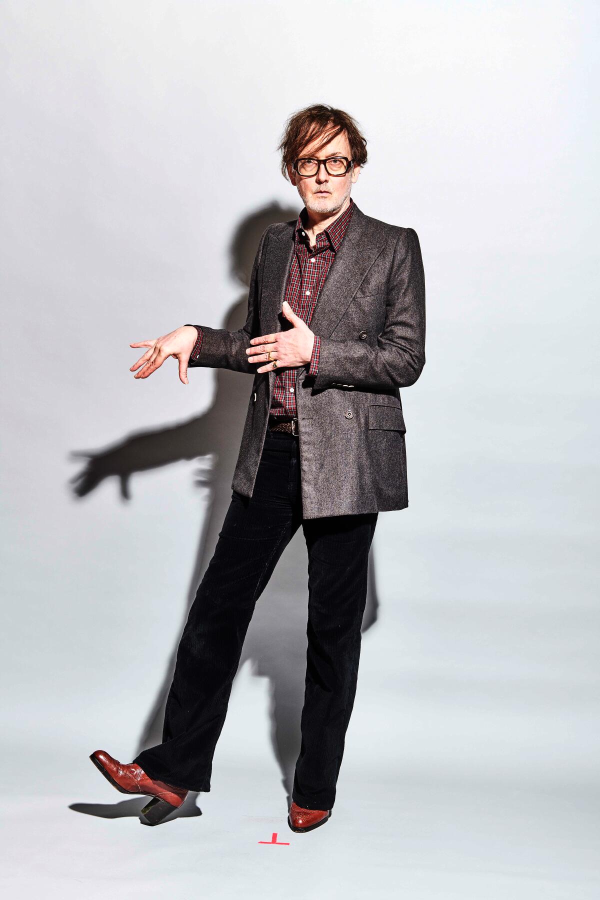 Jarvis Cocker of Pulp poses in a maroon shirt, grey blazer, black pants, and brown shoes.