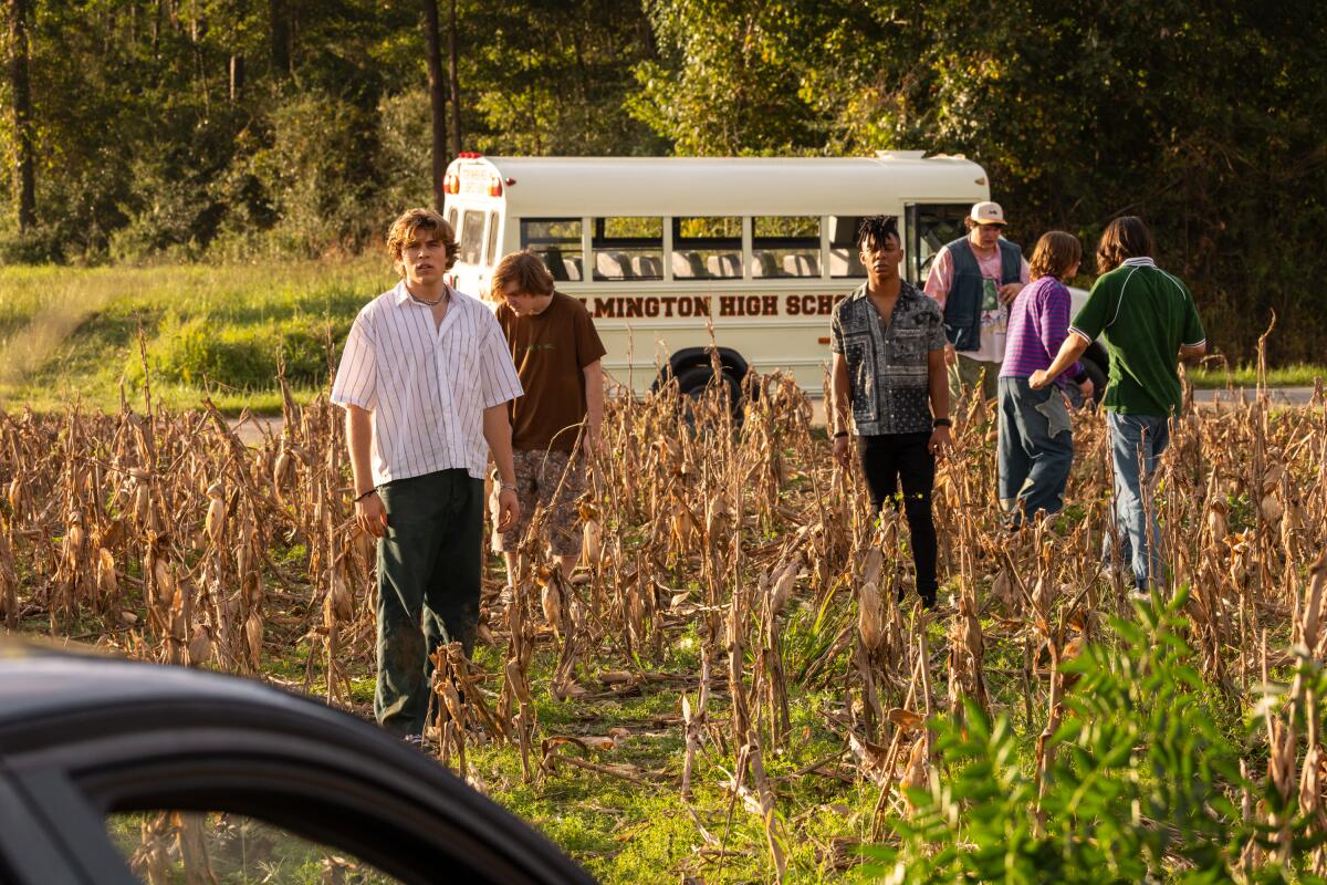 A group of teens walk through a dried corn field. A small white bus is parked on the road in the background.