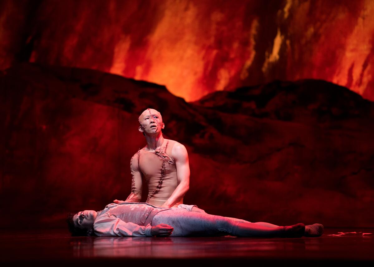 A dancer in monster makeup kneels beside another dancer on a stage with fiery backdrop.