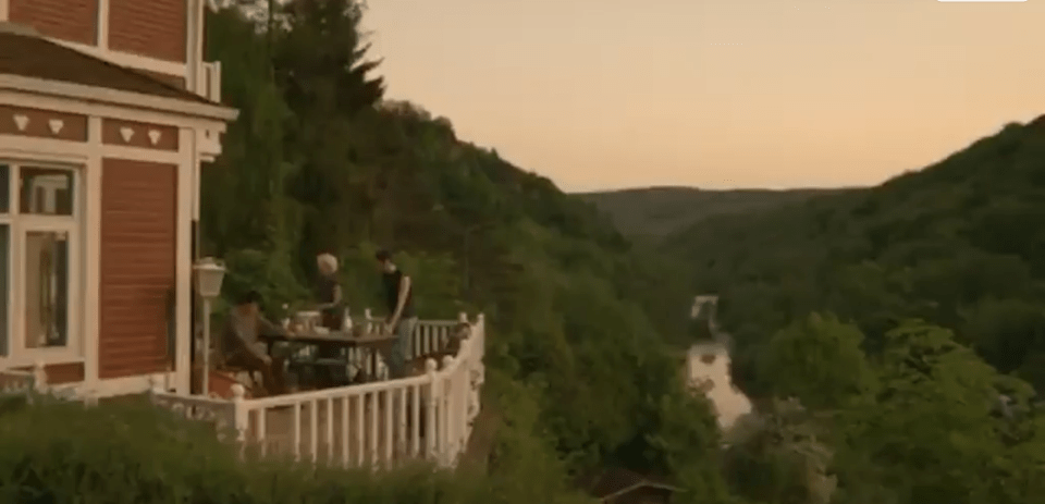 Three people enjoying drinks and food on a balcony overlooking a river and forested hills.