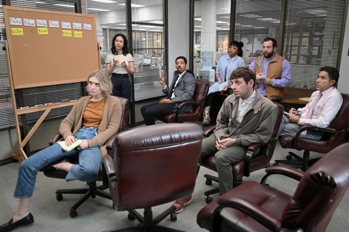 A group of people sitting on office chairs and on a table stand near a corkboard in glass-walled office.