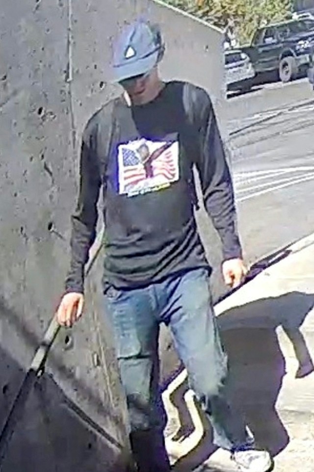 Surveillance image of a man wearing a blue hat and a black long-sleeve shirt with an American flag graphic.