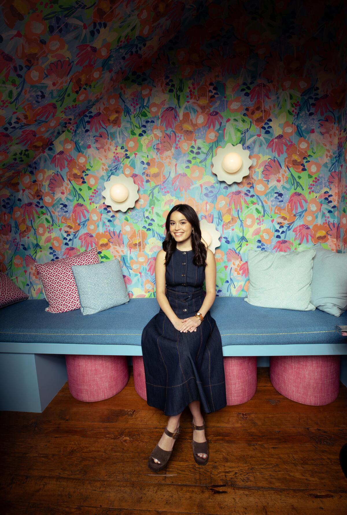 Annabelle Chang sits on a baby blue bench with blue and pink pillows in front of a wall with floral wallpaper.