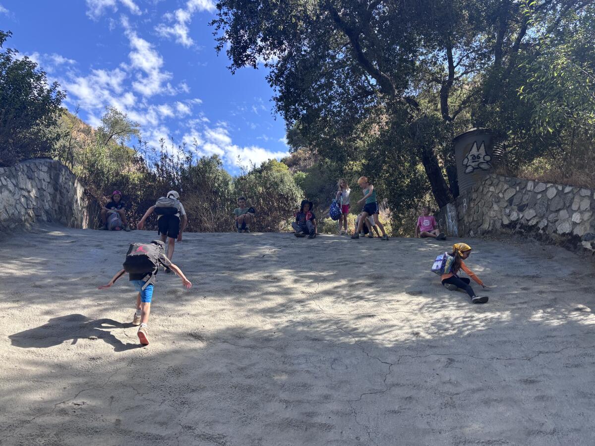 Children climb on a concrete drainage area, throwing backpacks down it and sliding down.