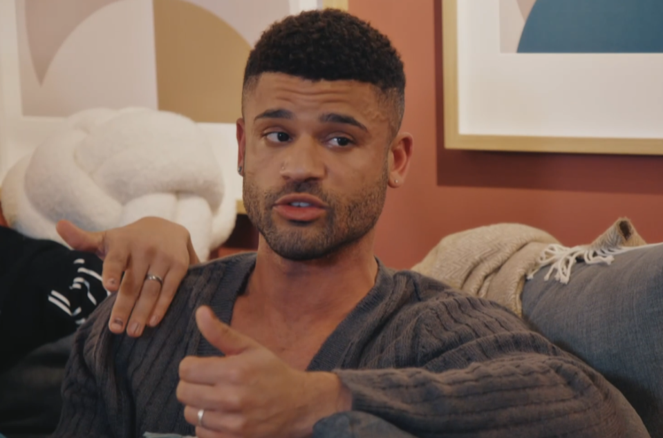 A man with short curly hair, stubble beard, and wearing a grey sweater, speaks while looking to his right.