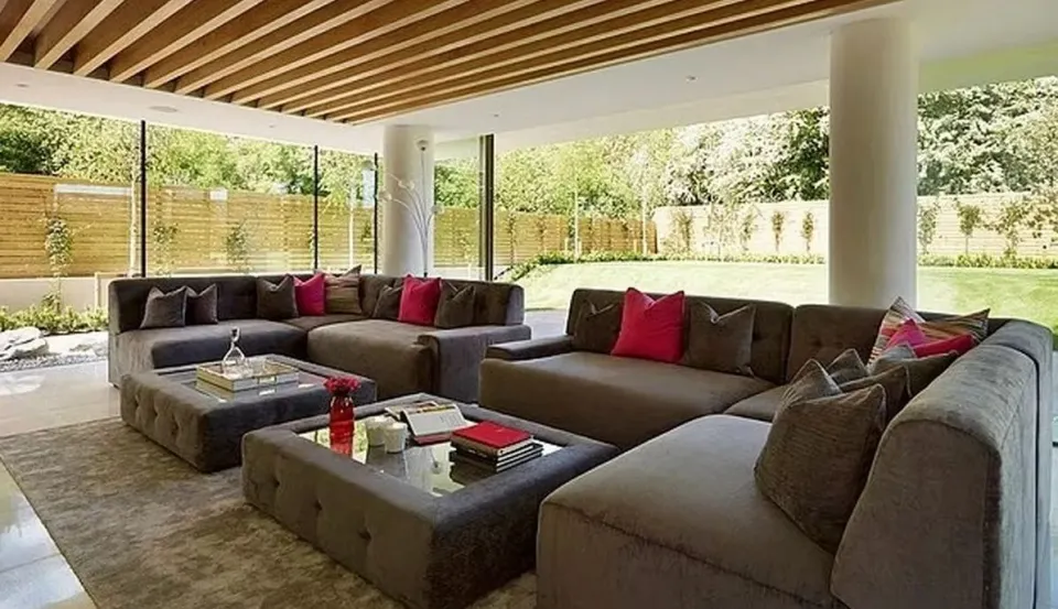 Cheryl Tweedy's former home in Hertfordshire, featuring a living room with multiple gray couches, glass coffee tables, and a view of a green garden.