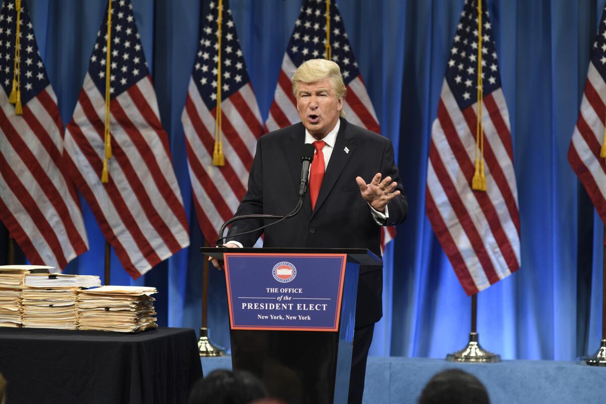 A man in a suit and red tie gesturing with his hand and standing behind a podium next to table stacked with folders.
