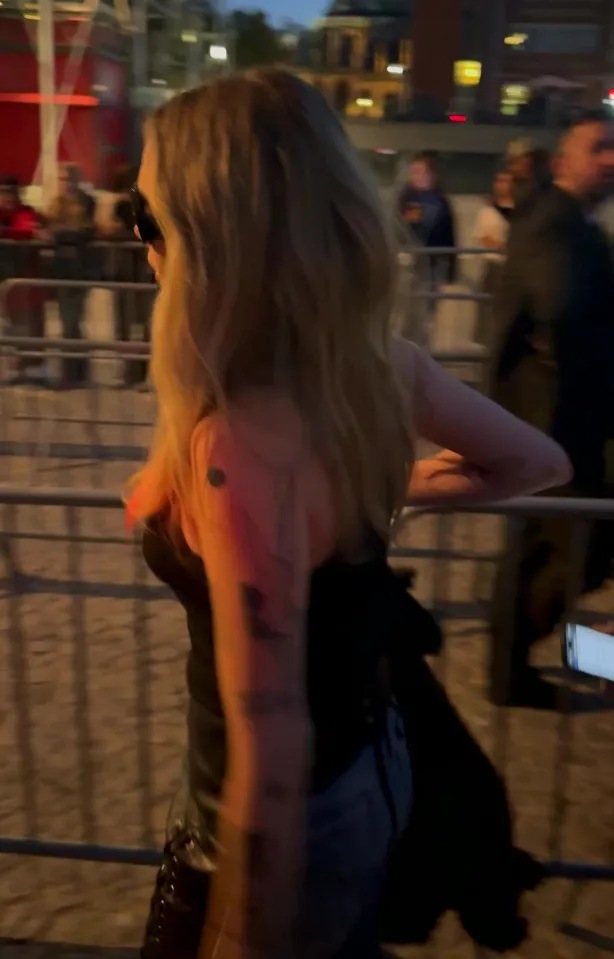 Cara Delevingne in a black top with long blonde hair, looking unhappy at a Stella McCartney fashion show.