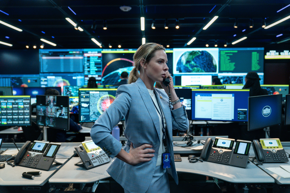 Olivia Walker in a light blue pantsuit talking on a black corded phone in a command center.
